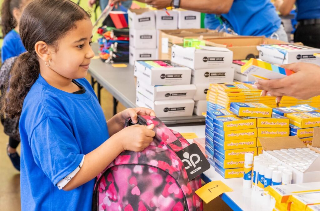 Una niña sostiene una mochila y mete en ella material escolar.